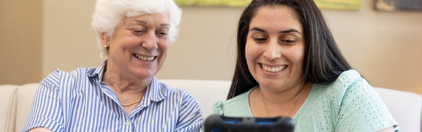 An elderly woman with a bright smile enjoys looking at a tablet with a young woman, illustrating a friendly interaction at Carlton Senior Living.
