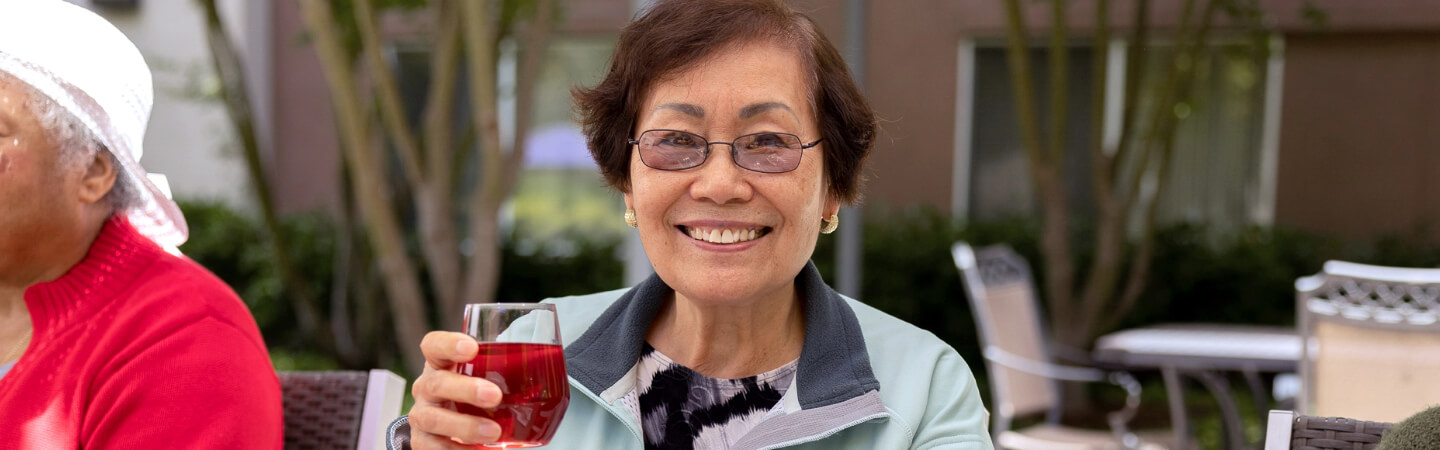 An elderly woman with glasses enjoys a beverage and outdoor social time at Carlton Senior Living.