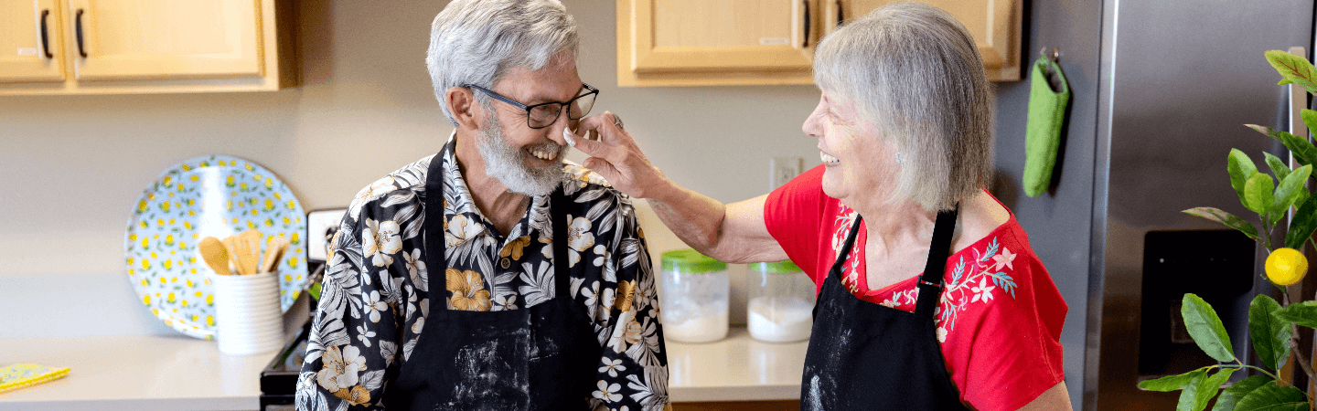 An elderly couple in a kitchen sharing a joyful moment, interacting with one another, both wearing aprons. This scene highlights the vibrant community life at Carlton Senior Living.