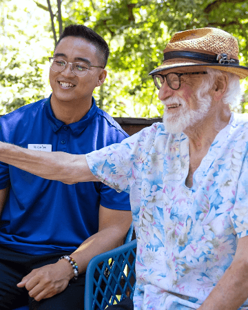 An elderly man with a straw hat and a floral shirt paints on a canvas, guided by a smiling Carlton team member in a blue uniform, exemplifying the engaging activities provided at Carlton Senior Living.