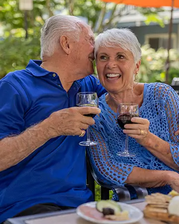 An elderly couple hold wine glasses and sit by a fountain, sharing a happy moment at Carlton Senior Living.