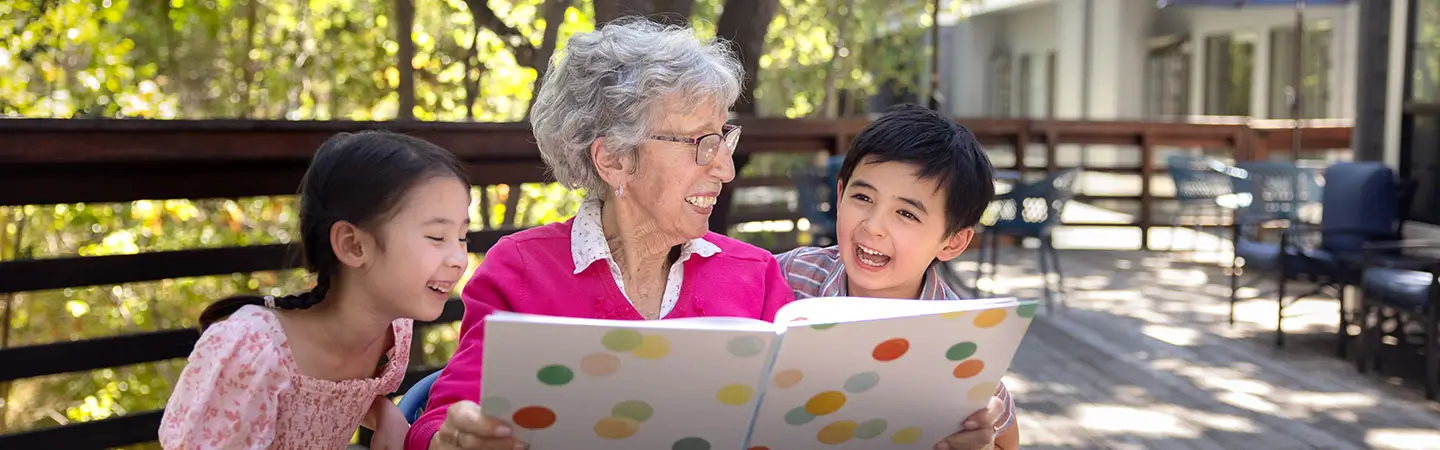 An elderly woman reads a book with two young children, a boy and a girl, who are laughing and sitting on a bench in an outdoor area, showing connections fostered at Carlton Senior Living.