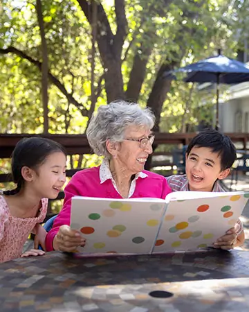 An elderly woman reads a book with two young children, a boy and a girl, who are laughing and sitting on a bench in an outdoor area, showing connections fostered at Carlton Senior Living.
