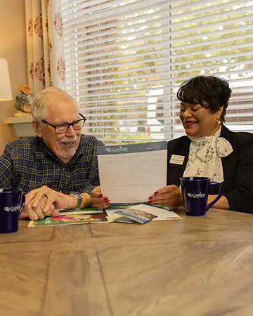 An elderly man in a plaid shirt and glasses laughs with a Carlton team member in a black and white outfit while reviewing a document together, sitting at a table with Carlton-branded mugs.