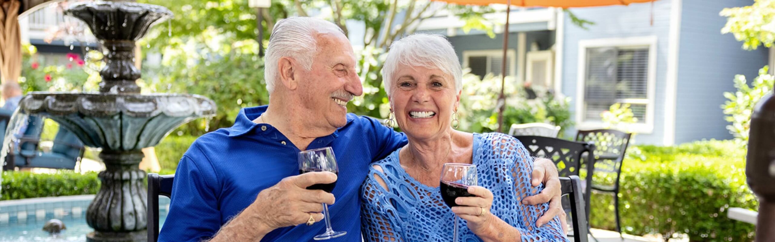 An elderly couple share a cheerful toast with glasses of red wine, seated by a fountain in a lush garden setting, expressing the joy and relaxation of life at Carlton Senior Living.