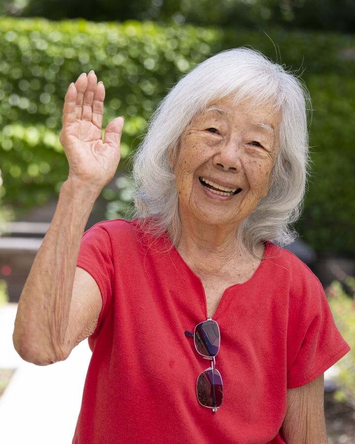 An elderly woman with silver hair and a radiant smile waves cheerfully in a garden, conveying the friendly community atmosphere at Carlton Senior Living.