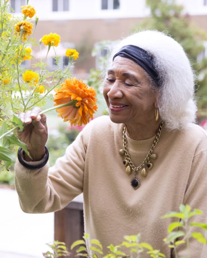 An elderly woman with white hair and a headband smells an orange flower in a lush garden, wearing a beige sweater and gold jewelry, illustrating the serene outdoor experiences available at Carlton Senior Living.