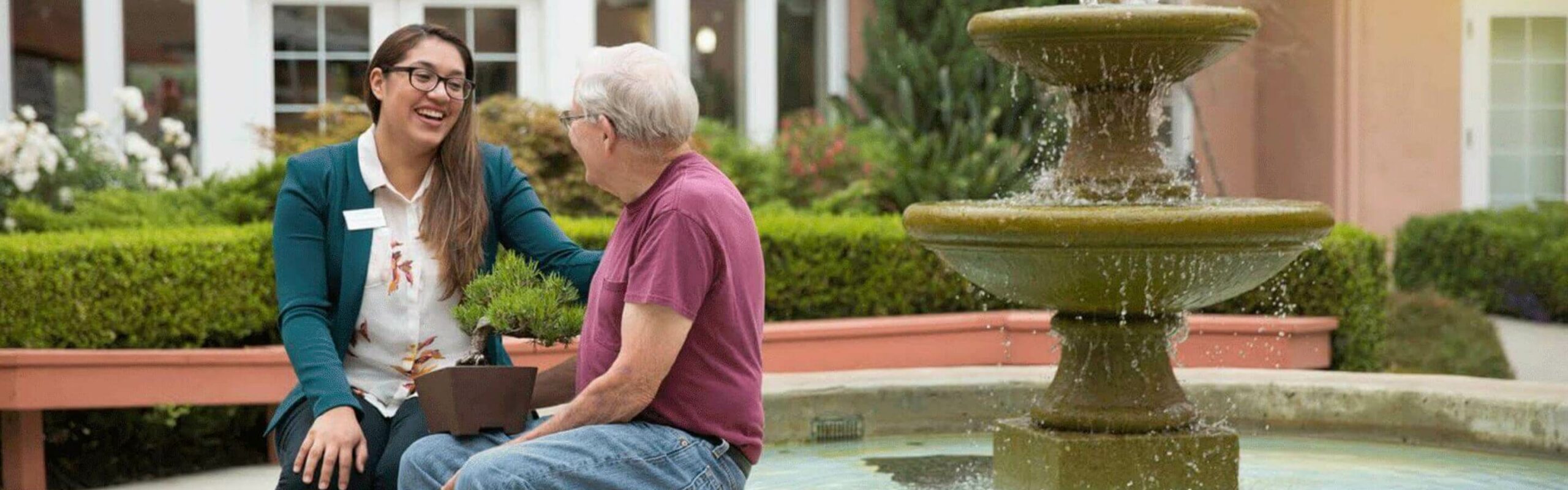 An elderly man and a Carlton team member share a laugh while seated on a bench by a fountain, representing the caring and friendly team interactions at Carlton Senior Living.