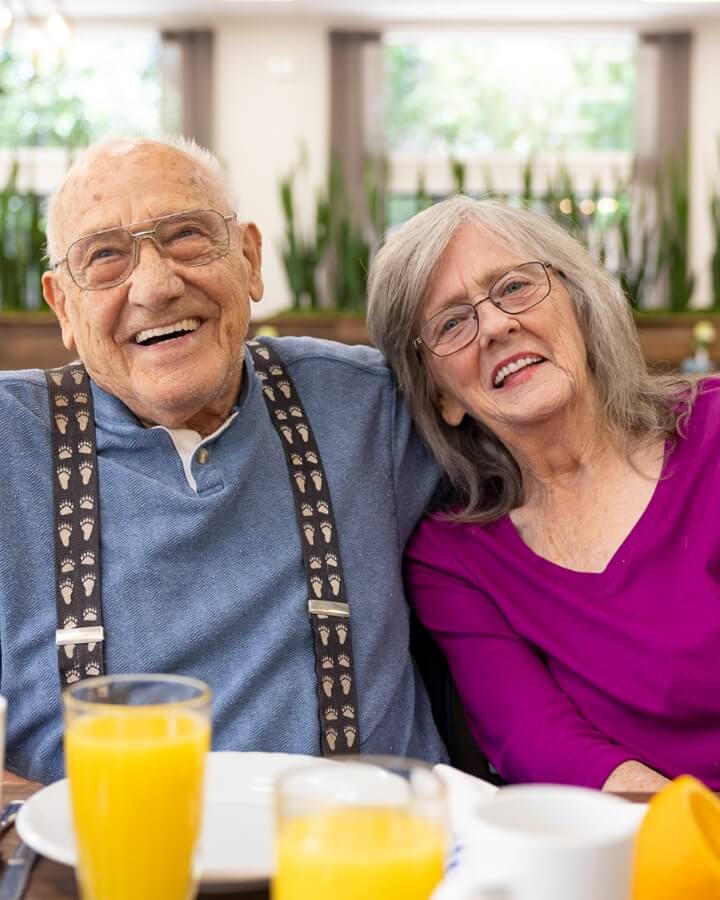 An elderly couple sitting together; the man is laughing and the woman leans on his shoulder smiling, demonstrating the community atmosphere at Carlton Senior Living.