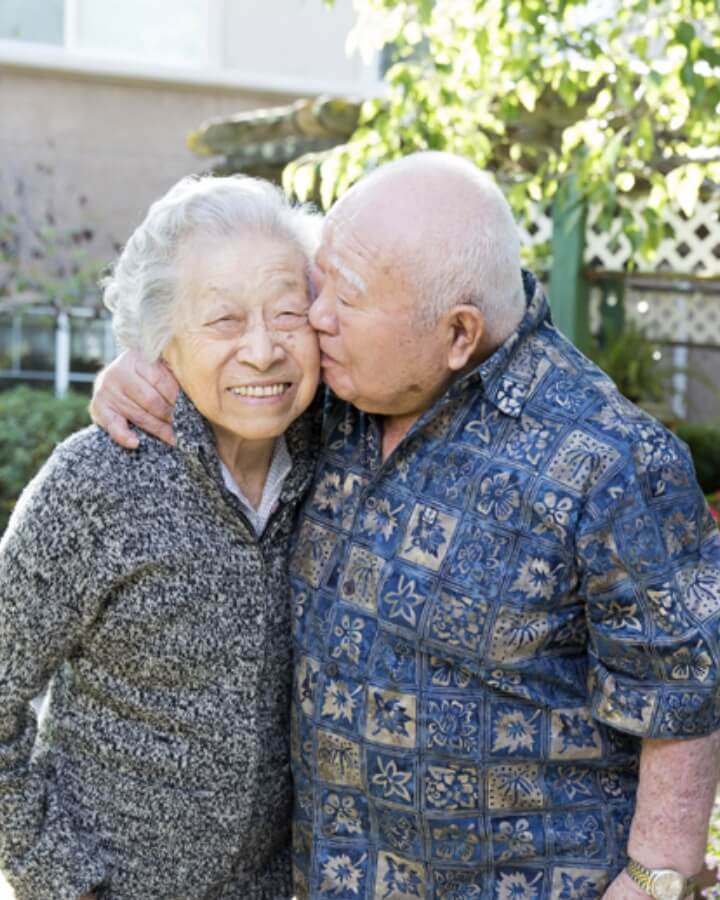 An elderly couple, with the man giving a kiss on the cheek to a woman, both smiling in a garden at Carlton Senior Living.