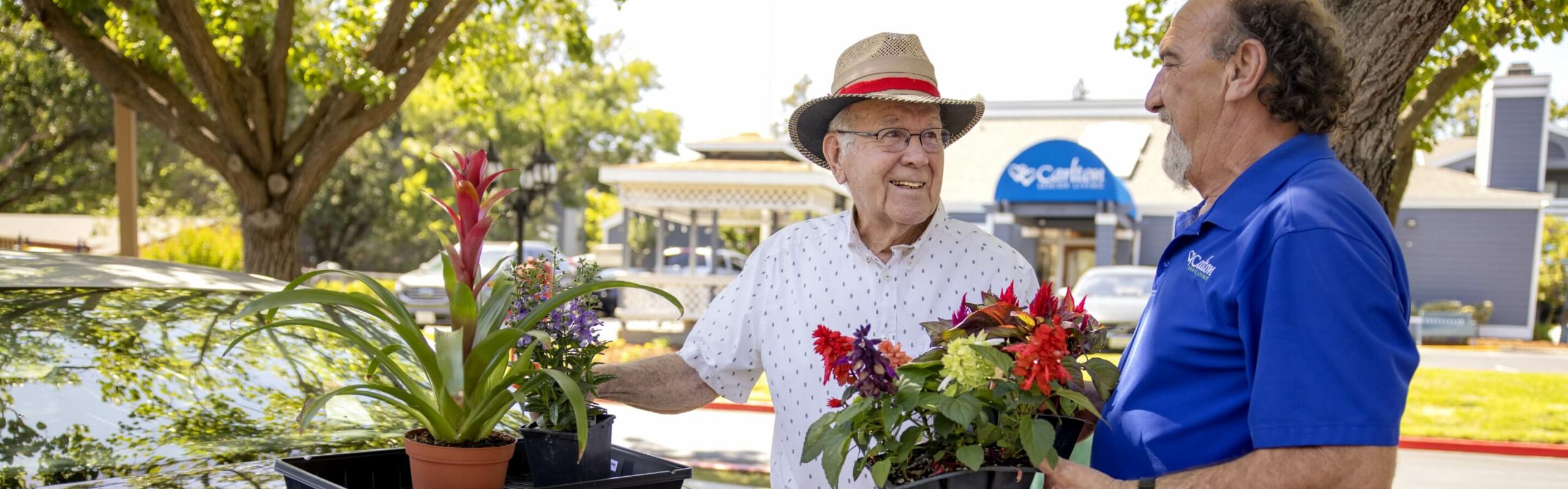 An elderly man in a hat and white shirt smiling by colorful plants accompanied by a Carlton team member in a blue shirt, showcasing the community's outdoor setting.