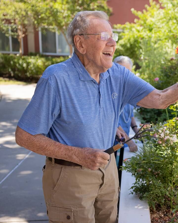 An elderly man in a blue shirt laughs and enjoys flowers in a blooming garden portraying outdoor life at Carlton Senior Living.