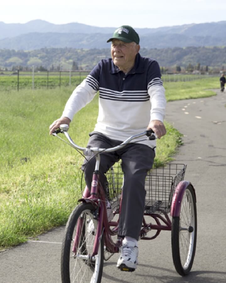 An elderly man enjoys a bicycle ride along a winding road, admiring green fields and mountain views, depicting an active lifestyle at Carlton Senior Living.
