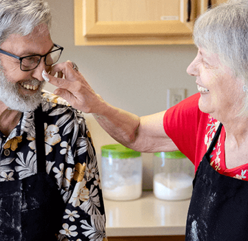 An elderly couple in a kitchen sharing a joyful moment, interacting with one another, both wearing aprons. This scene highlights the vibrant community life at Carlton Senior Living.