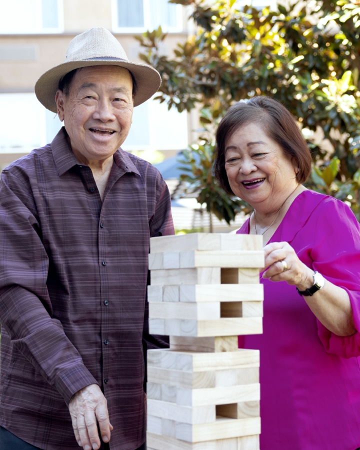 An elderly couple laugh together while playing a game outdoors, with the man in a hat and striped shirt and the woman in a fuchsia top, embodying the active and social lifestyle at Carlton Senior Living.